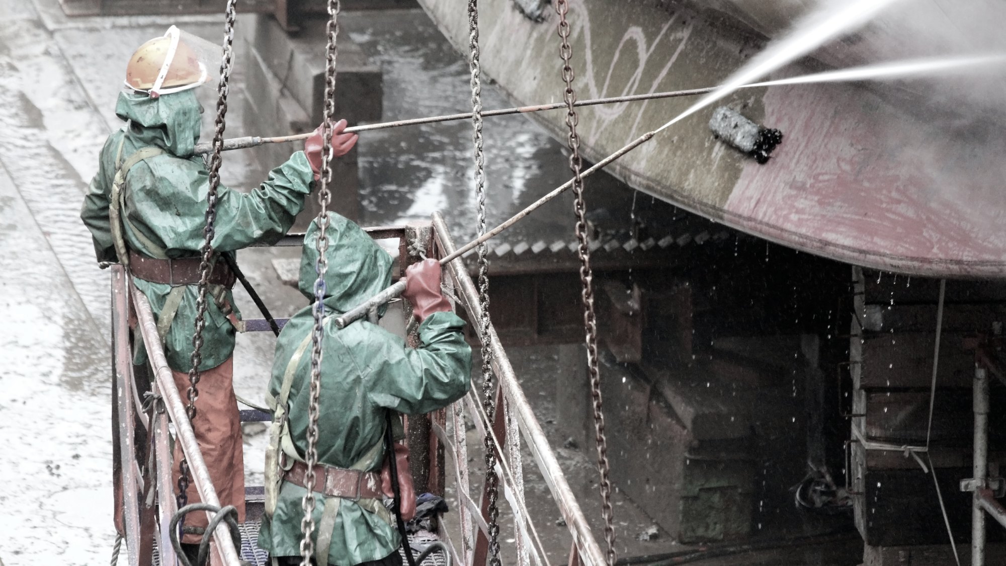 Two workers in protective suits pressure-washing the hull of a ship in drydock, suspended on a cradle