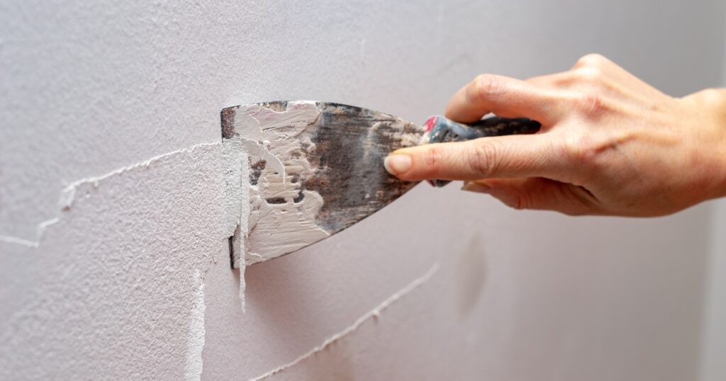 A hand scraping cracked paint and filler from a wall with a putty knife